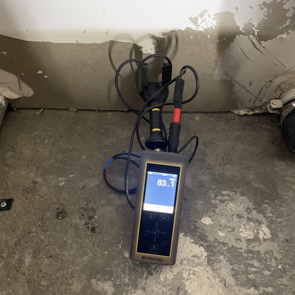 Feuchtemessgerät mit Elektroden auf dem Estrich vor einer aufgestemmten Wand, die Anzeige zeigt einen Messwert von 83,1